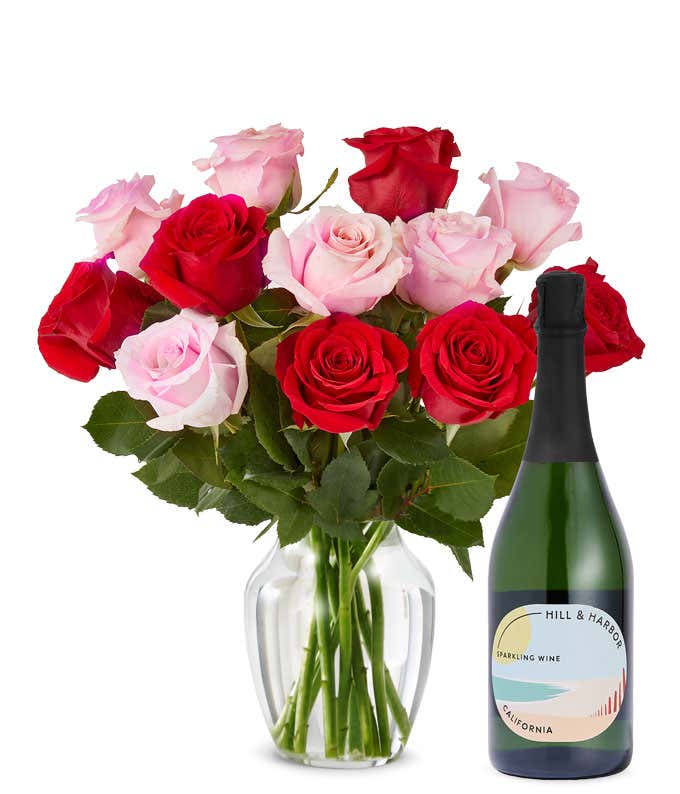 A mix bouquet of one dozen red and blush roses fills a clear glass vase and is paired with a bottle of Hill & Harbor California Sparkling Wine.
