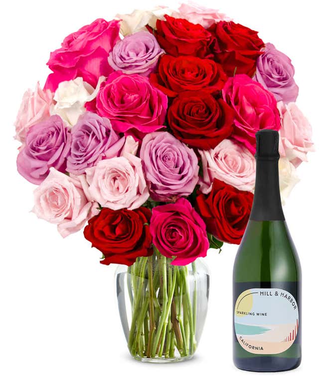 Two dozen pink, purple and red roses arranged in a clear glass vase sits beside a bottle of Hill &amp; Harbor California Sparkling Wine.