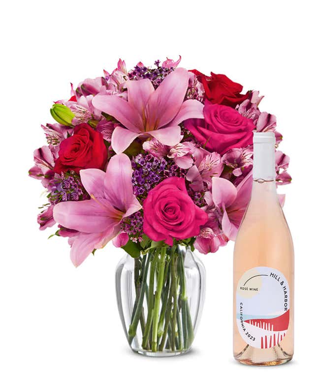 A bouquet of pink lilies, pink roses, and greenery arranged in a clear glass vase sitting next to a bottle of Hill &amp; Harbor California Ros&eacute;.