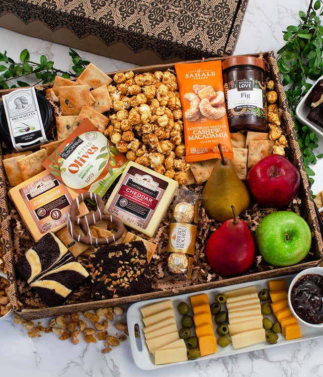 Gift basket with assorted cheeses, crackers, olives, fresh apples and pears, caramel popcorn, fig spread, nut mix, chocolate treats, and pastries arranged in a decorative box with a small cheese board display.