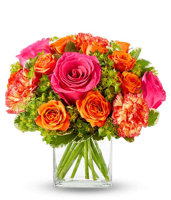 An arrangement of hot pink roses, orange spray roses, orange carnations, bupleurum, lily grass, and pittosporum, all beautifully arranged in a clear glass cube vase.