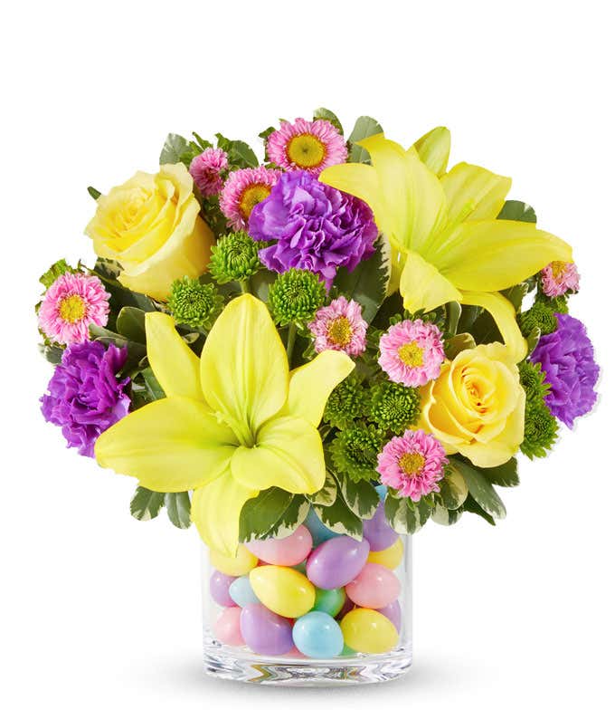 Yellow Asiatic lilies, yellow roses, purple carnations, pink Matsumoto asters, and green button poms arranged in a clear glass vase filled with colorful pastel Easter eggs